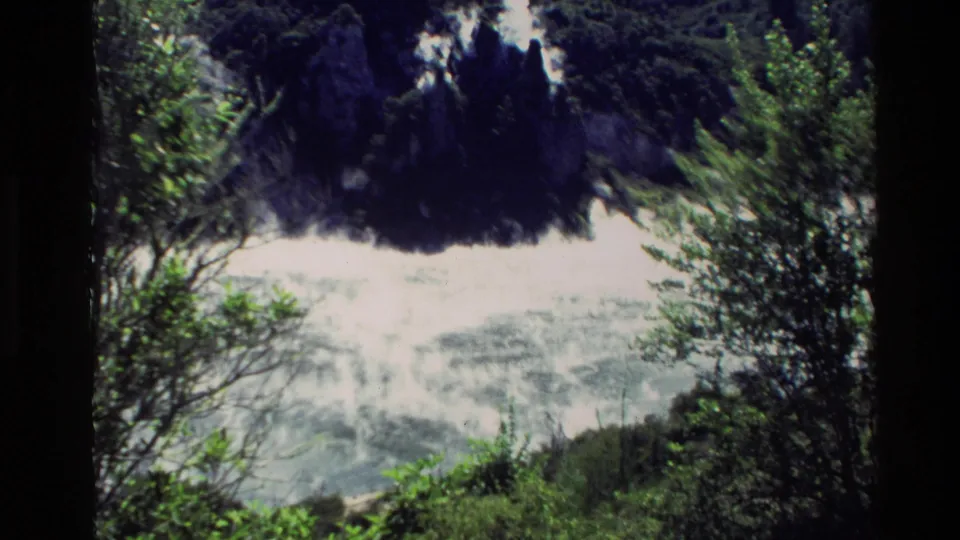 1981 New Zealand 8mm frame showing pale volcanic steam against a dark rocky hillside with green foliage in the foreground.