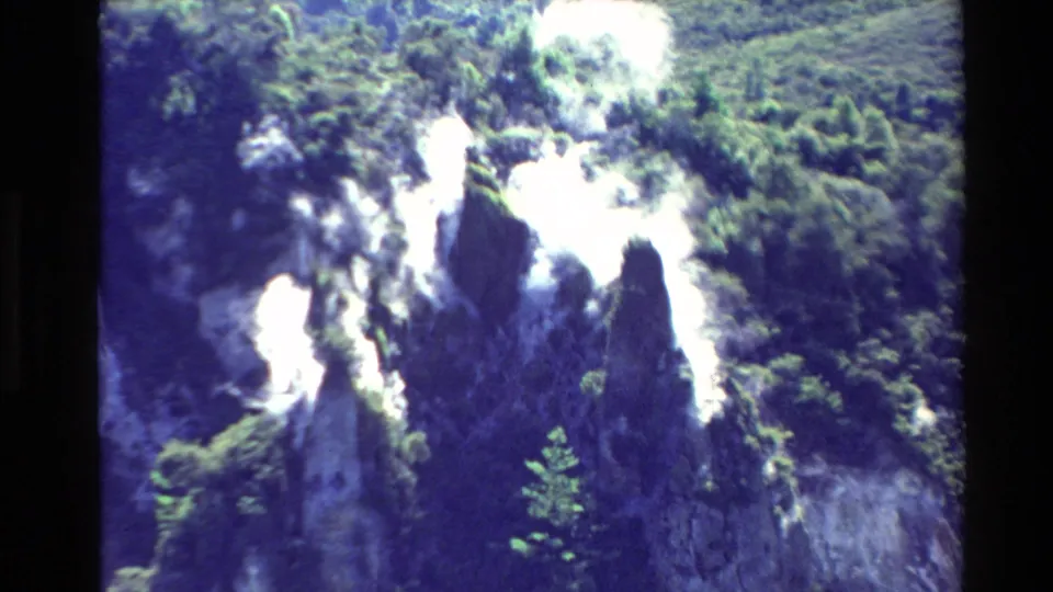 1981 New Zealand 8mm frame showing tall volcanic steam columns rising through shadowed rock and scrub-covered hillside.