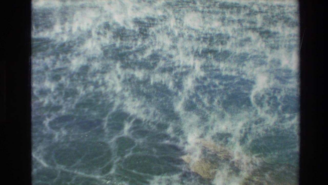 Foaming ocean water churns around dark rocks in a grainy 8mm frame.