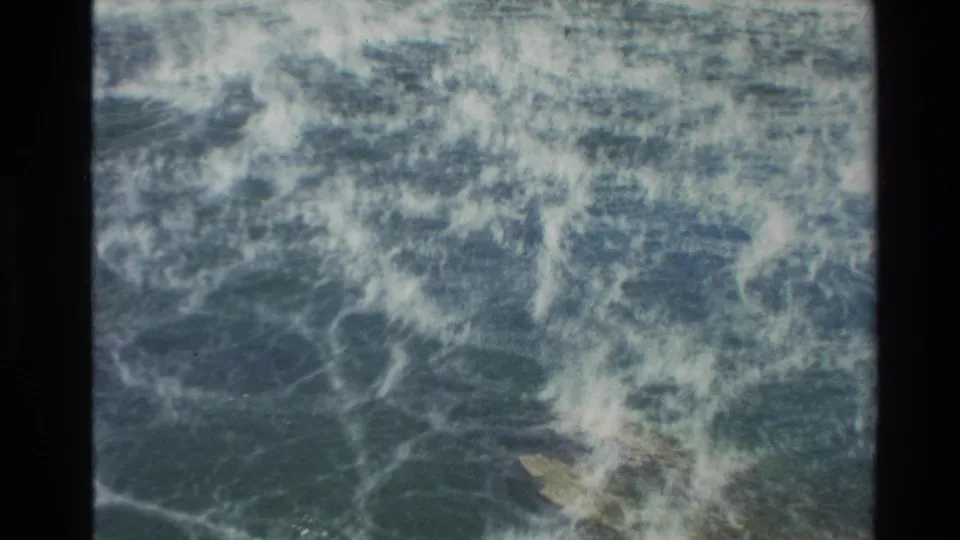 Archival 8mm footage from New Zealand shows turbulent surf washing across rocks, with no people visible.