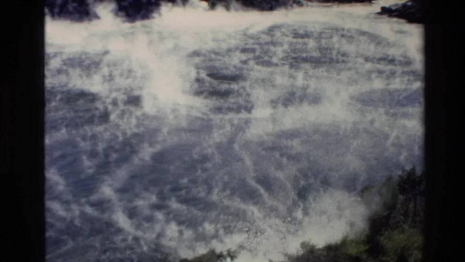 New Zealand 8mm footage shows churning surf near rocks, with an indistinct submerged form partly obscured by foam.