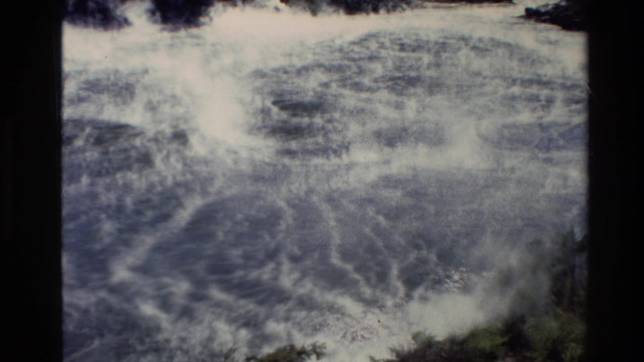 1981 New Zealand 8mm frame showing dense steam or mist over shallow blue-green coastal water near a rocky, vegetated shore.