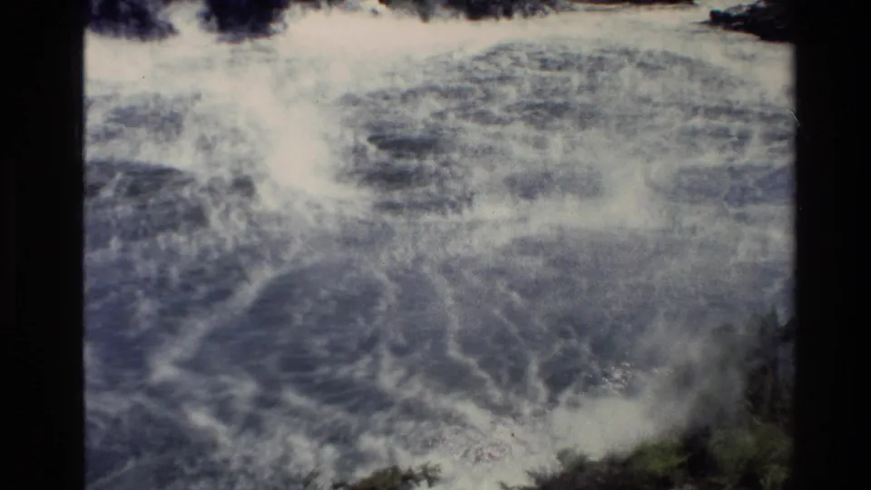1981 New Zealand 8mm frame showing dense steam or mist over shallow blue-green coastal water near a rocky, vegetated shore.