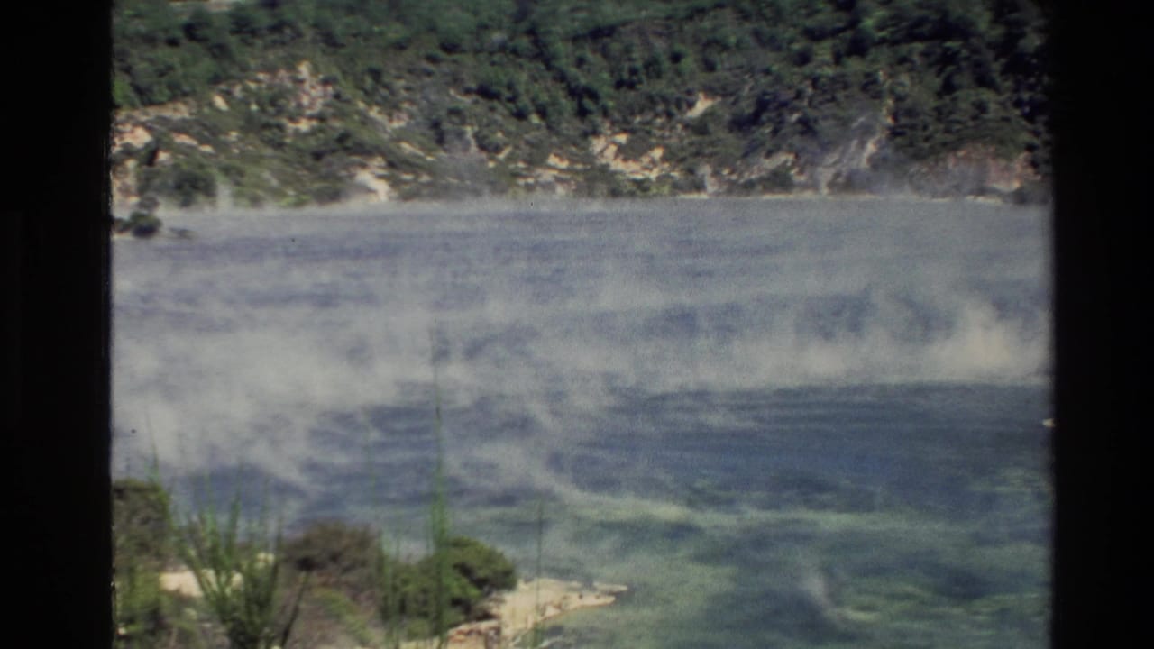 A blue-green cove with steam or mist rising below a wooded hillside.