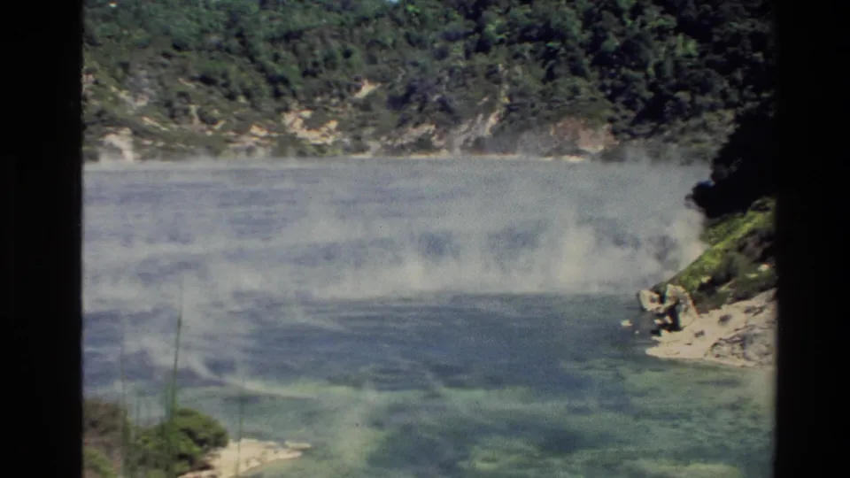 1981 New Zealand 8mm frame looking across a shallow cove with vapor on the water and a rocky, tree-covered shoreline beyond.