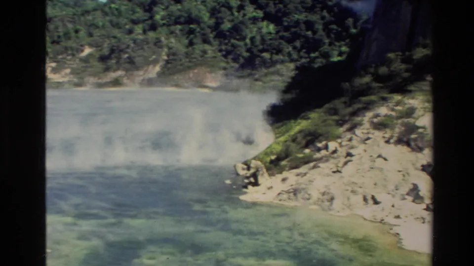 1981 New Zealand 8mm frame showing vapor over shallow blue-green water beside a pale rocky beach and vegetated coastal slope.
