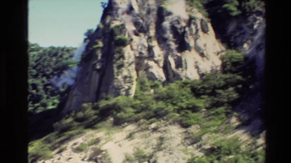 1981 New Zealand home-movie frame of a steep rock face with vegetation and steam rising through a geothermal valley.