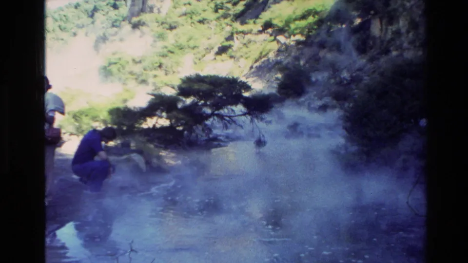 1981 New Zealand home-movie frame with distant visitors near steaming geothermal water beneath trees and rocky slopes.