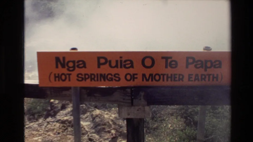 Orange sign reading Nga Puia O Te Papa, with English text for hot springs of Mother Earth, beside a steamy New Zealand geothermal area.