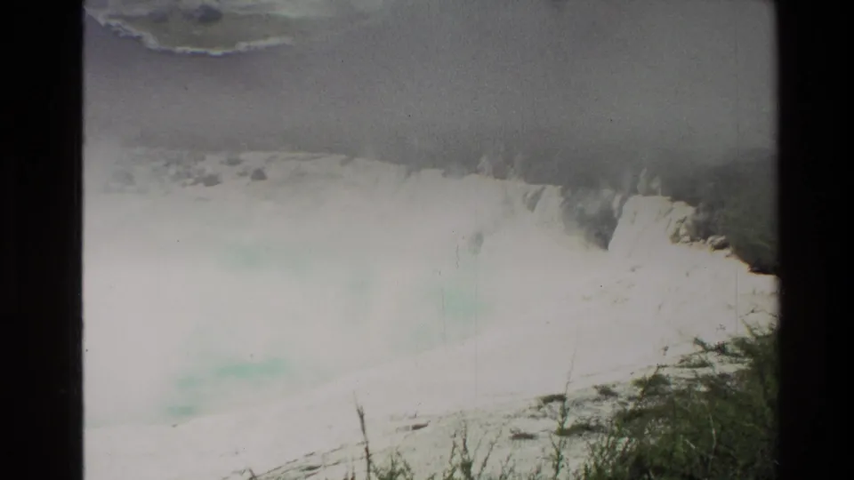 A pale blue steaming geothermal pool in New Zealand appears beside light mineral crust and scrub in faded 8mm footage.