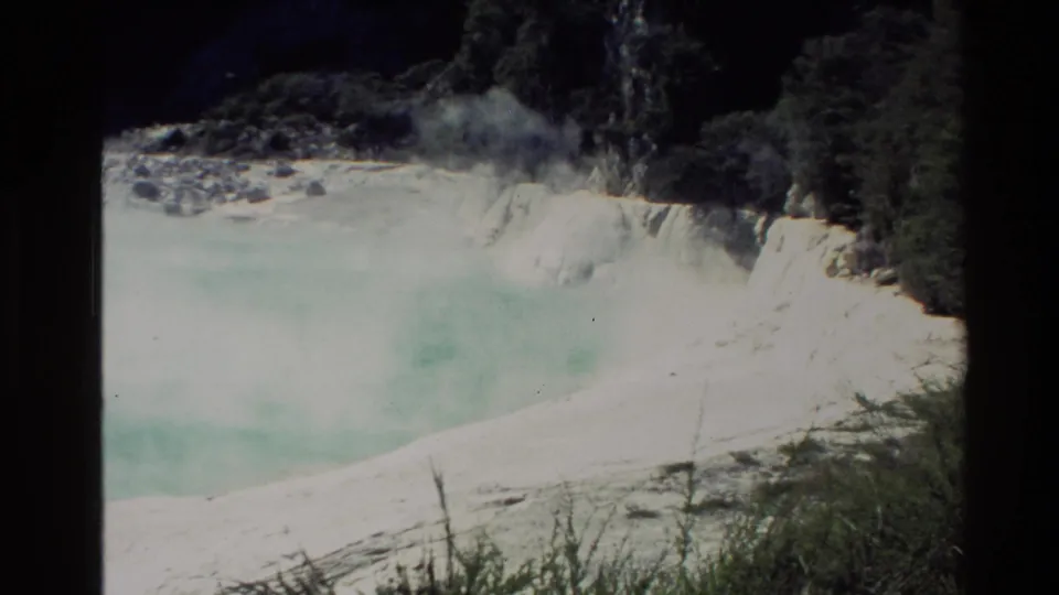 1981 New Zealand home-movie frame of a steaming geothermal pool with dark rocks, scrub, and pale mineral edges.