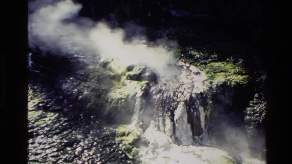 1981 New Zealand 8mm frame with steam drifting from a rocky geothermal vent beside reflective water.