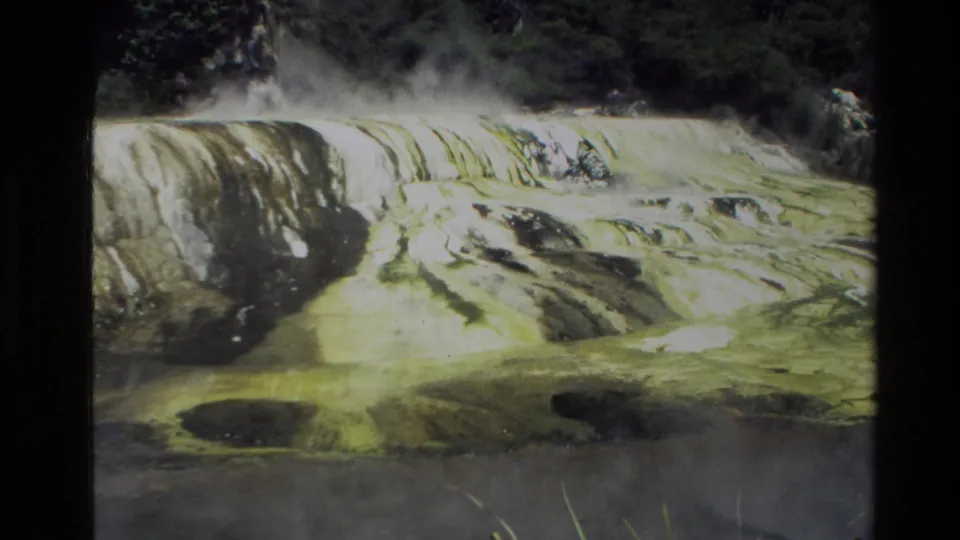 Archival 8mm view from New Zealand in 1981 of steaming pale terrace-like formations with green and dark vegetation around them.