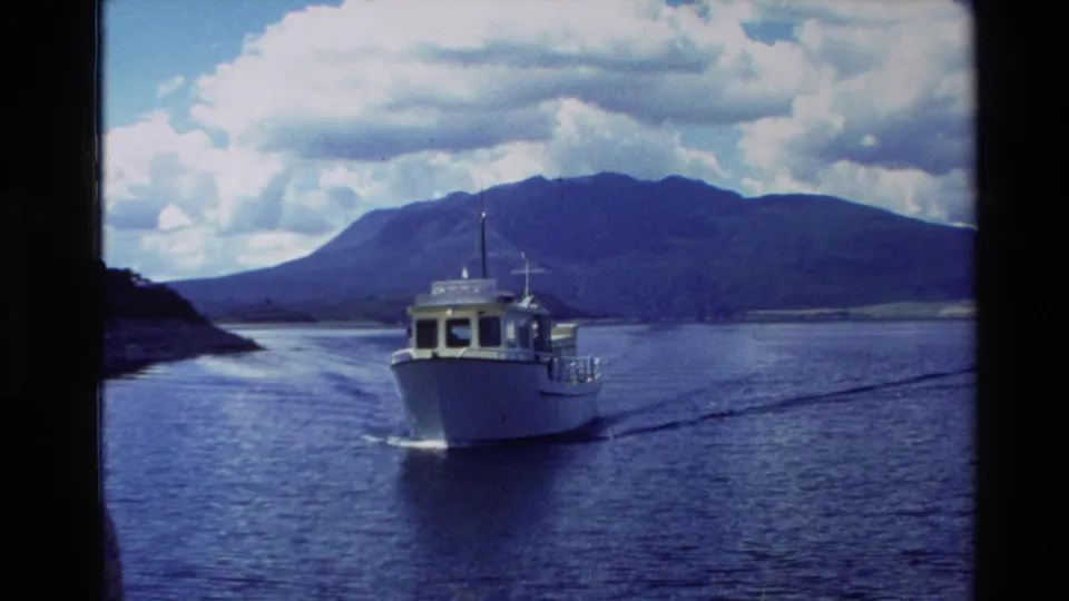 New Zealand 1981 home-movie frame of a white motorboat crossing a calm waterway against a dark mountain and bright clouds.