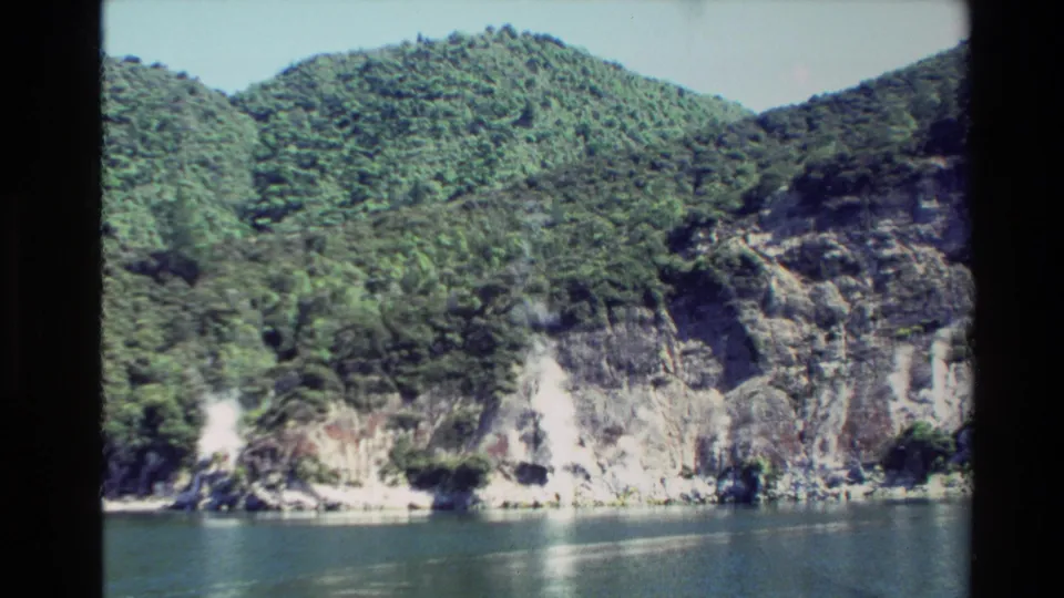 New Zealand shoreline with dense green hills, pale rocky cliffs, and calm water in a vintage 8mm travel frame.