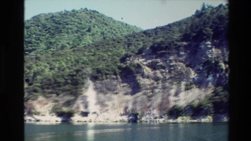 1981 New Zealand home-movie view of a forested slope, steep rock face, and quiet water from a moving vantage point.