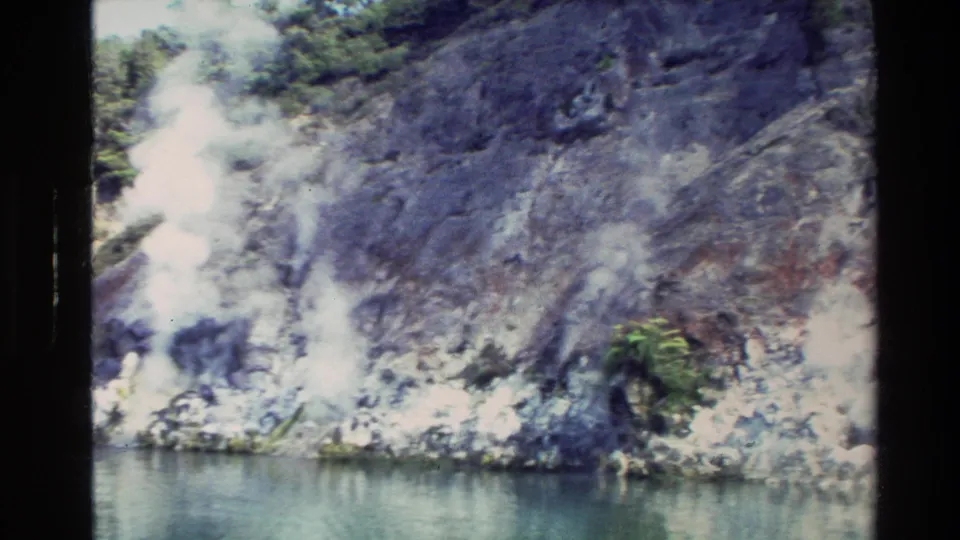 Vintage 8mm frame of a steep rocky New Zealand shoreline where pale steam or mist appears near calm water.