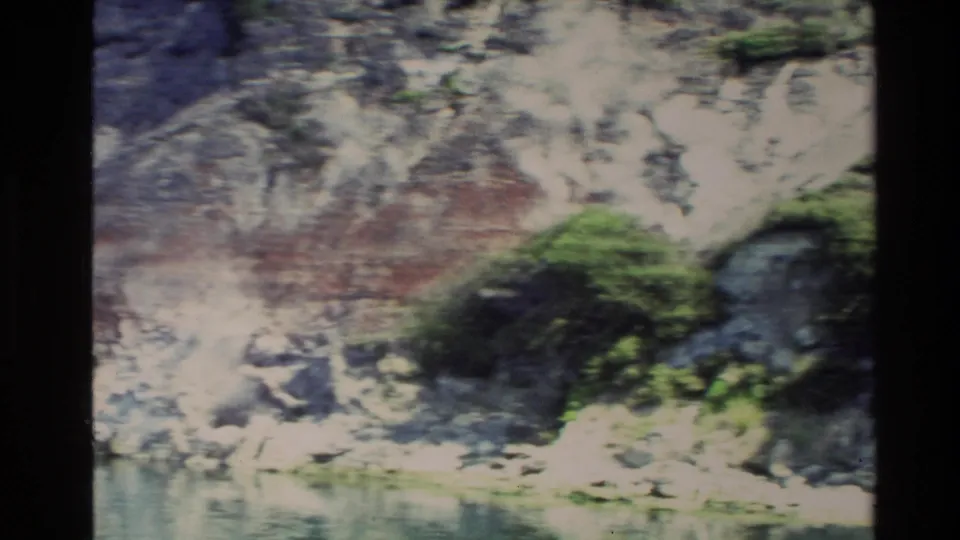 1981 New Zealand 8mm frame of pale rock, mossy growth, and still water along a riverbank.