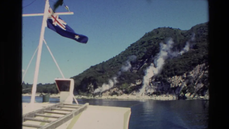 1981 New Zealand travel footage from a boat, with the national flag on a mast and steam rising from a forested lakeside hill.