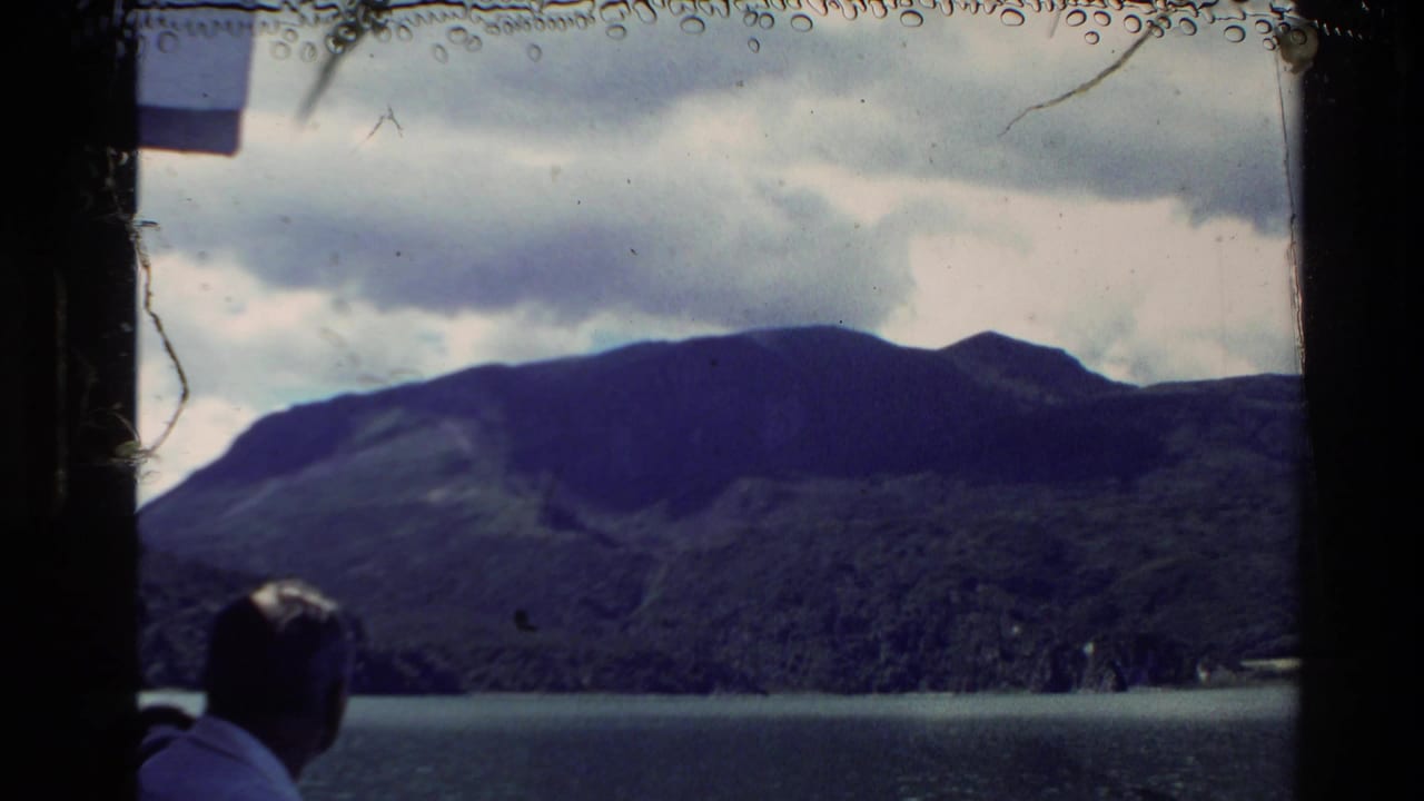 New Zealand 1981 8mm frame with a man viewed from behind facing a mountain range across the lake, framed by dark film edges.