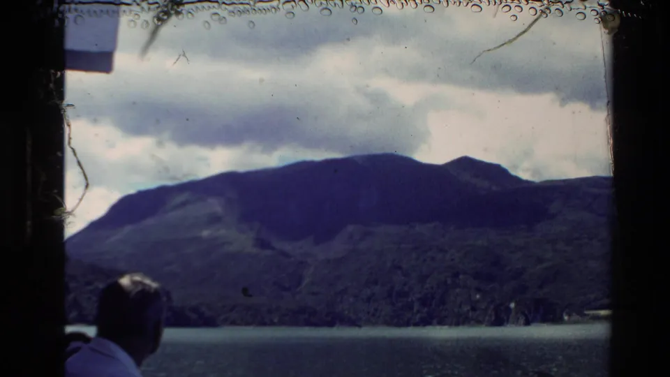 New Zealand 1981 8mm frame with a man viewed from behind facing a mountain range across the lake, framed by dark film edges.