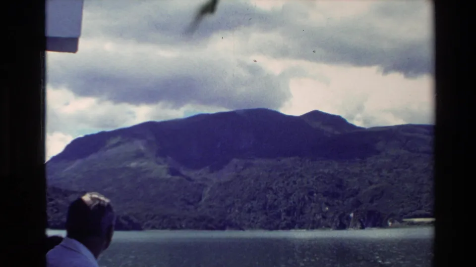 New Zealand 1981 home-movie view of a man looking over a lake toward green mountain slopes and a cloud-filled sky.