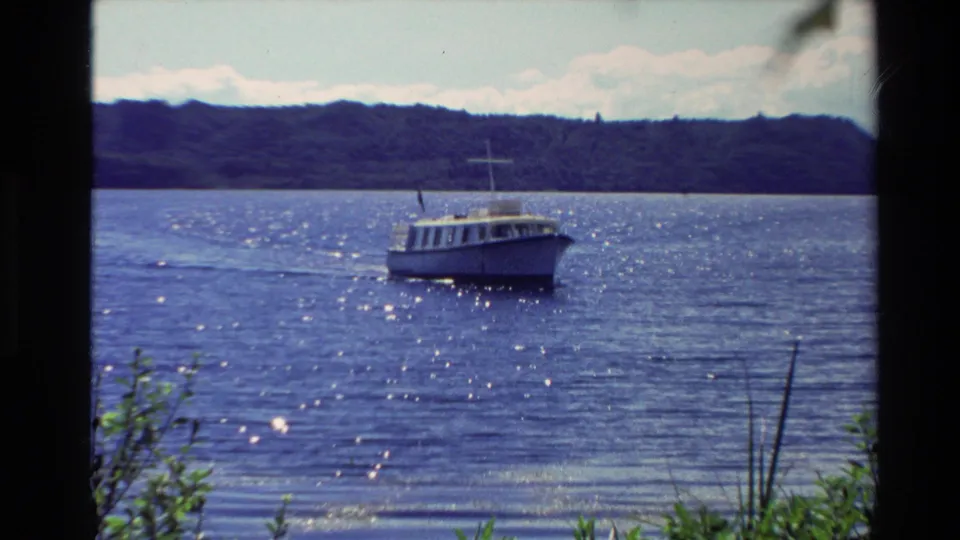 New Zealand 1981 8mm frame showing a small motorboat on sunlit lake water, with low hills and foreground shoreline plants.