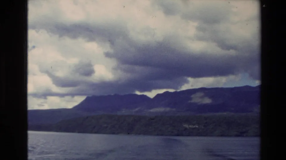 1981 New Zealand 8mm frame showing a broad water view with dark forested shoreline, distant mountains, and heavy cloud cover.