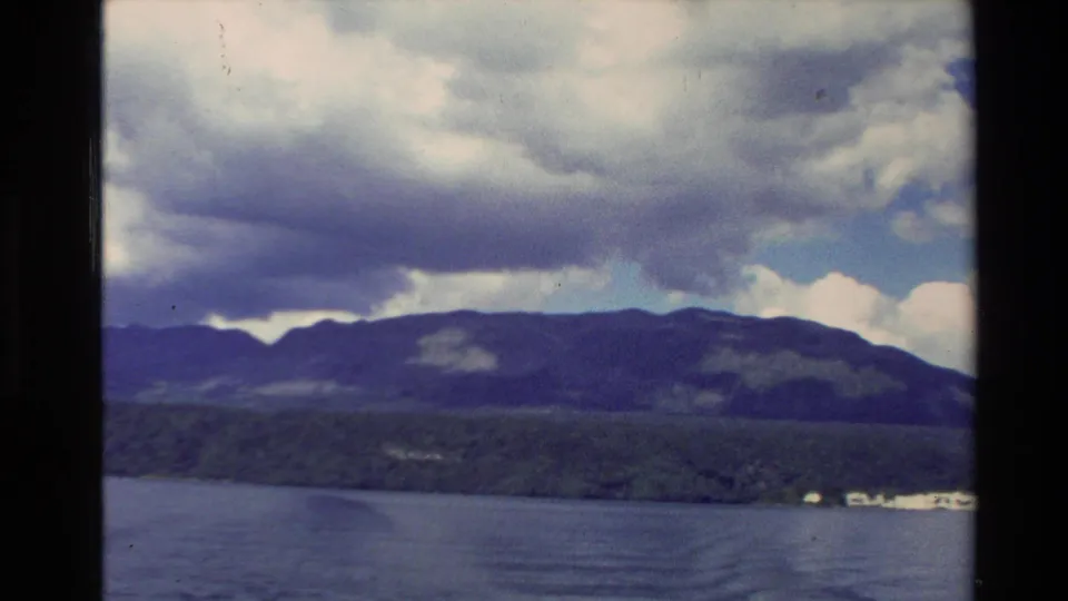 1981 New Zealand home-movie frame with blue water in the foreground, a wooded shoreline, and mountains beneath broad clouds.