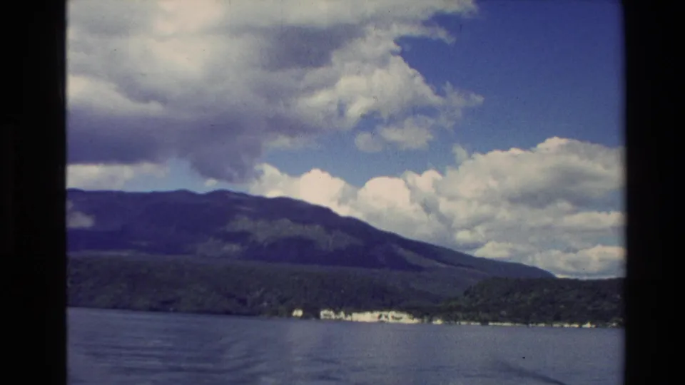 Vintage 8mm New Zealand travel view of lake water, mountain ridge, distant shore, and bright cloud formations.