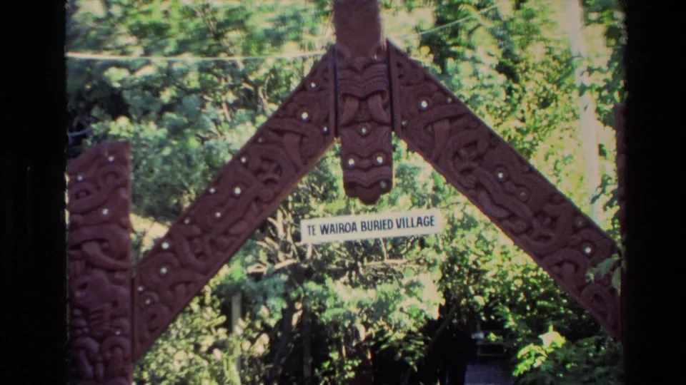 New Zealand home-movie frame showing a carved wooden entrance and a readable Te Wairoa Buried Village sign among trees.