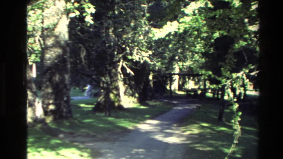 1981 New Zealand 8mm frame showing a shaded walkway, mature trees, lawn, and small distant visitors at the heritage site.