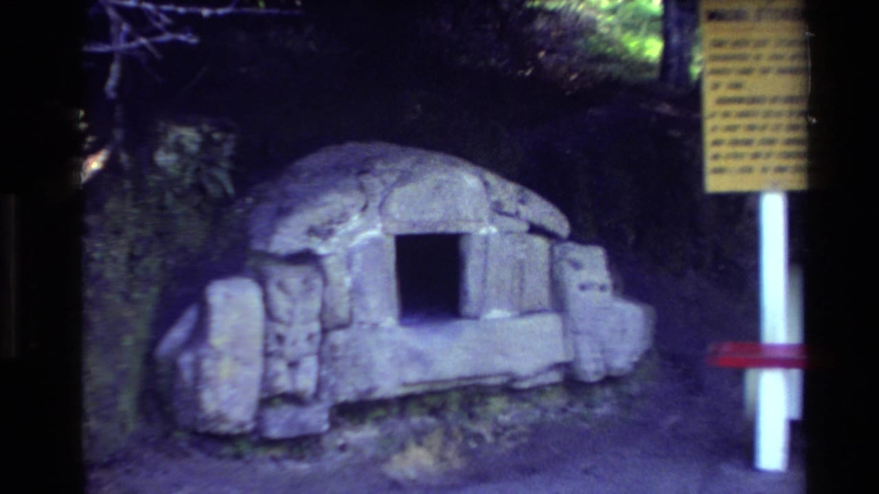 Stone hut facade with a dark rectangular opening, yellow sign, red bench, and trees behind it.