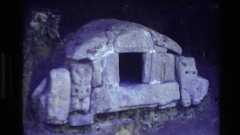 1981 New Zealand frame with a closer handheld view of a stone Māori-style hut facade and dark rectangular opening among trees.