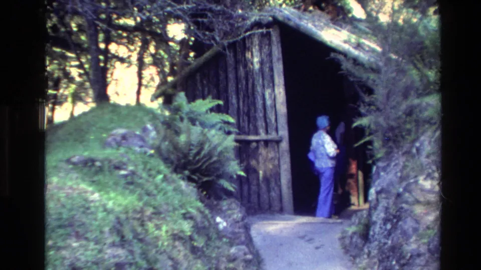1981 New Zealand home-movie frame showing a woman at the entrance of a rustic timber and thatched structure surrounded by ferns.