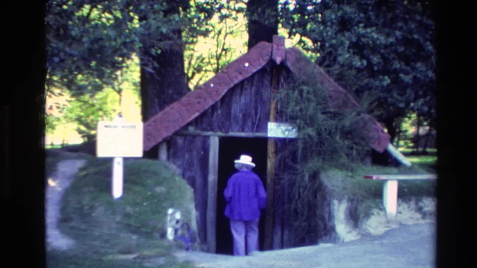 1981 New Zealand frame showing a woman in a purple jacket and white hat entering a Māori-style thatched wooden structure with signs nearby.