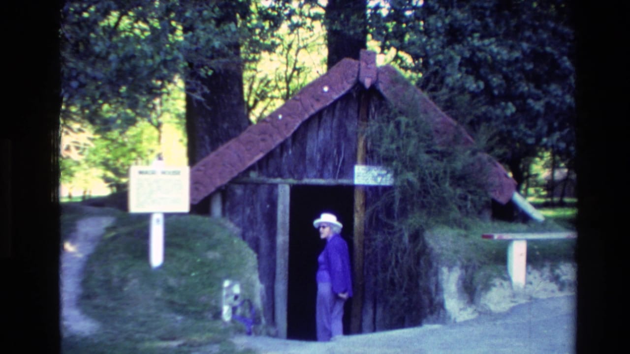 Tourist in a purple jacket standing beside a rustic wooden hut and white site sign.