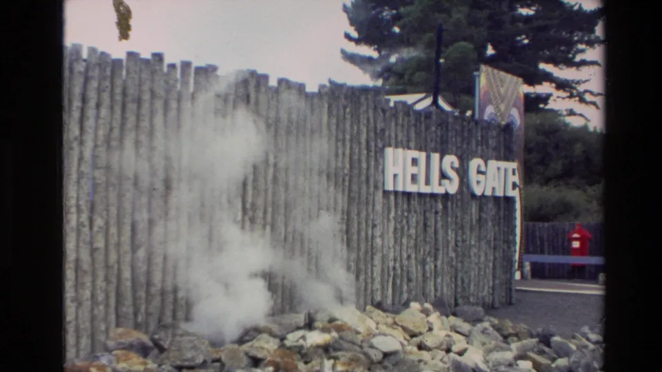 1981 New Zealand travel frame showing the readable Hells Gate sign on a wooden fence with geothermal steam rising across rocks.
