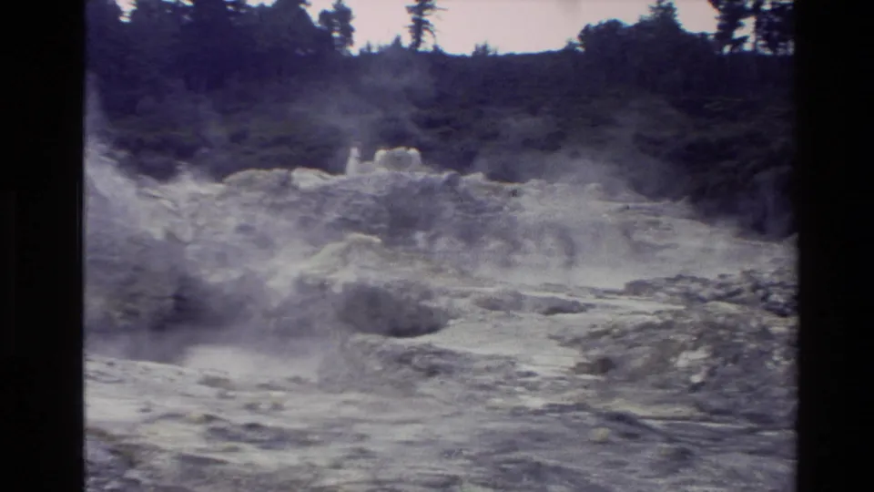 1981 New Zealand home-movie frame of a steaming geothermal landscape with rocky bubbling ground and wooded hills behind it.