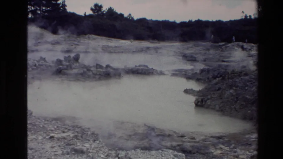 Wider New Zealand 1981 8mm frame showing a milky geothermal pool, rocky mineral banks, drifting steam, and distant trees.