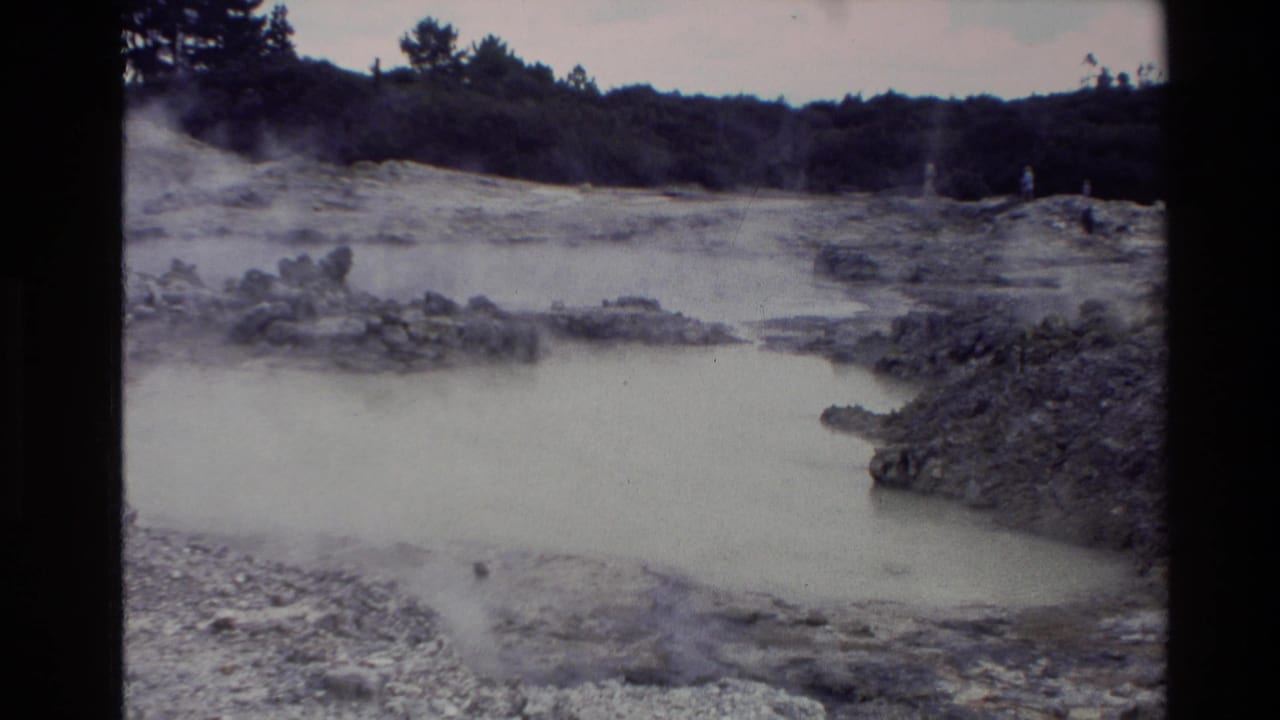 1981 New Zealand 8mm frame showing a steaming geothermal pool, pale mineral terrain, dark vegetation, and small distant visitors.