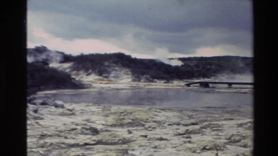 1981 New Zealand 8mm frame showing an overcast geothermal basin with water, low hills, and a distant wooden bridge.