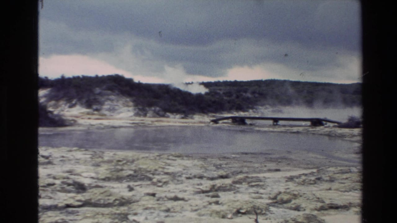 Wide view of a steaming geothermal area with a wooden walkway, pale ground, low hills, and cloudy sky.