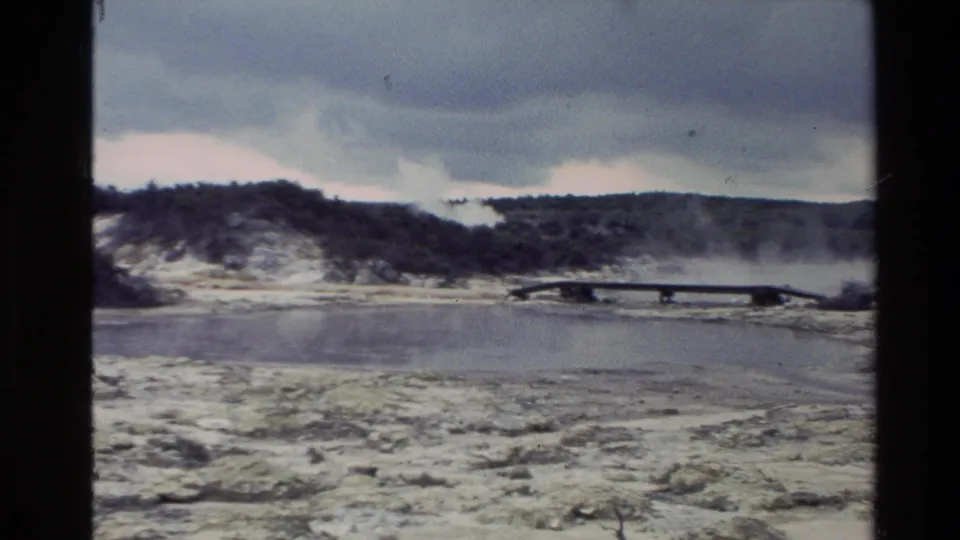 1981 New Zealand 8mm frame showing a steaming geothermal landscape, wooden walkway, mineral-streaked ground, low hills, and overcast sky.