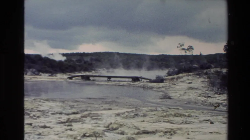 1981 New Zealand 8mm frame with a wooden walkway crossing a steaming geothermal field and pale mineral ground under cloud cover.