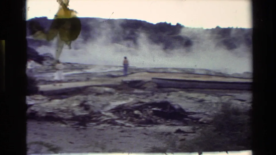1981 New Zealand frame with a distant visitor, raised boardwalks, steam clouds, pale ground, and wooded hills behind.