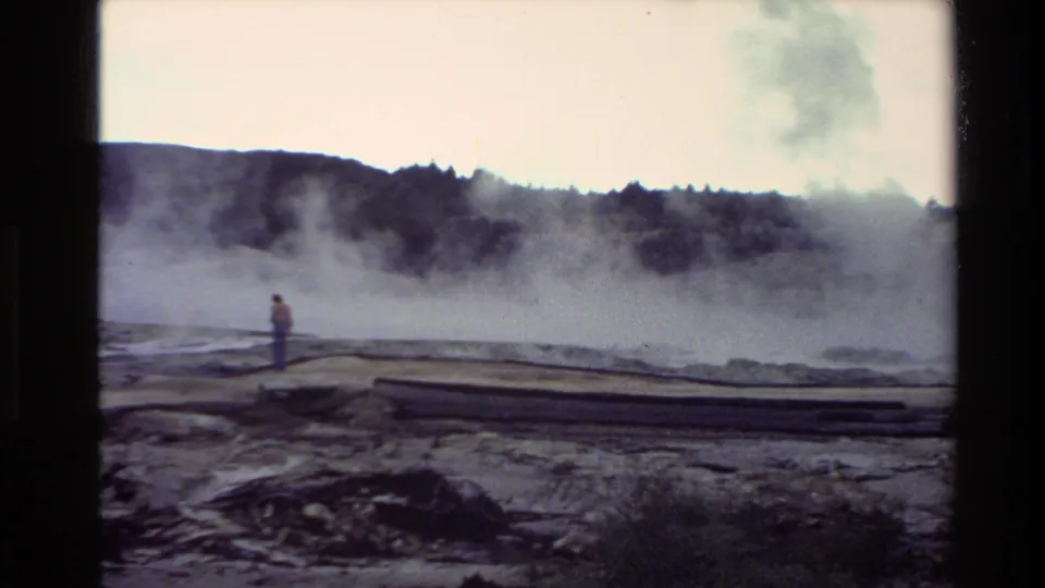 New Zealand 1981 8mm frame showing a visitor near boardwalks with heavy geothermal steam and dark hills in the background.