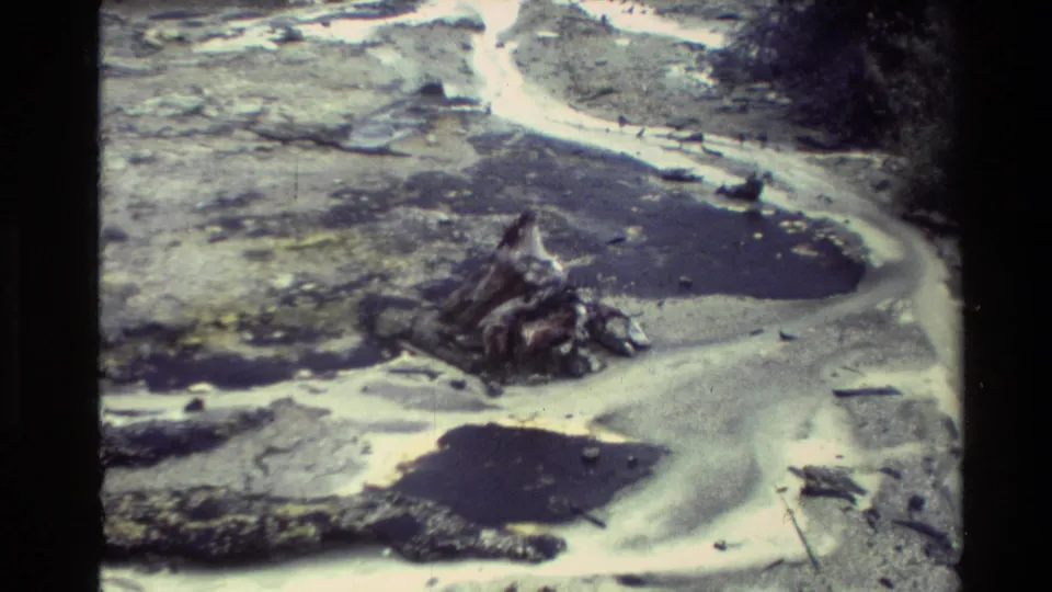 1981 New Zealand home-movie frame showing a winding runoff channel, mineral deposits, dark pools, rocks, and geothermal terrain.