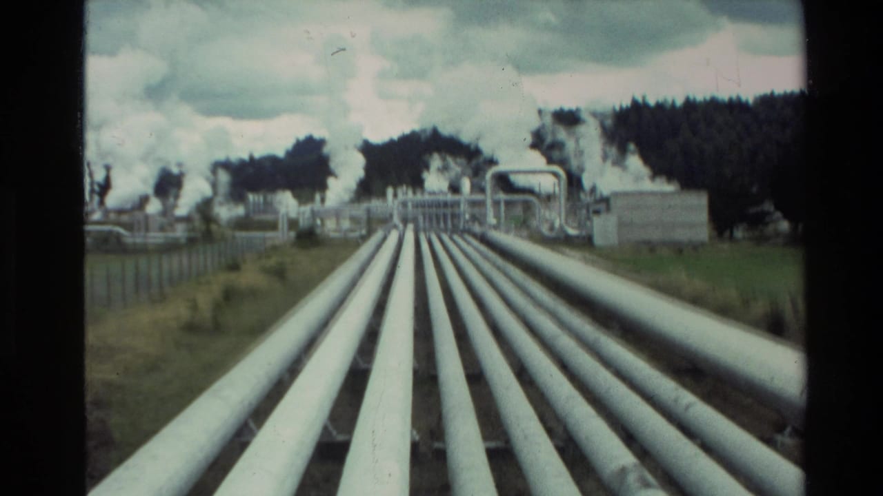 1981 New Zealand 8mm view of large white geothermal pipes running toward industrial structures with steam plumes beyond.