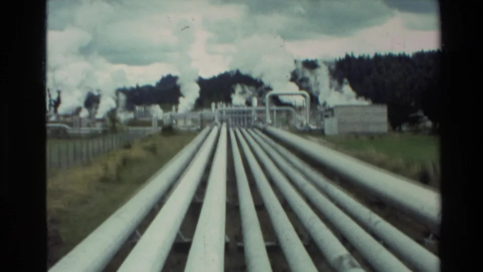 1981 New Zealand 8mm view of large white geothermal pipes running toward industrial structures with steam plumes beyond.